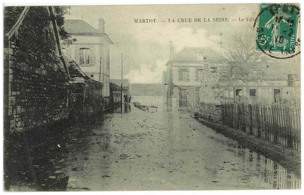Carte postale 1910. Martot. La crue de la Seine envahie le village. Des personnages observent, devant une passerelle installée sur la route submergée. Source : Collection Archives Départementales 27
