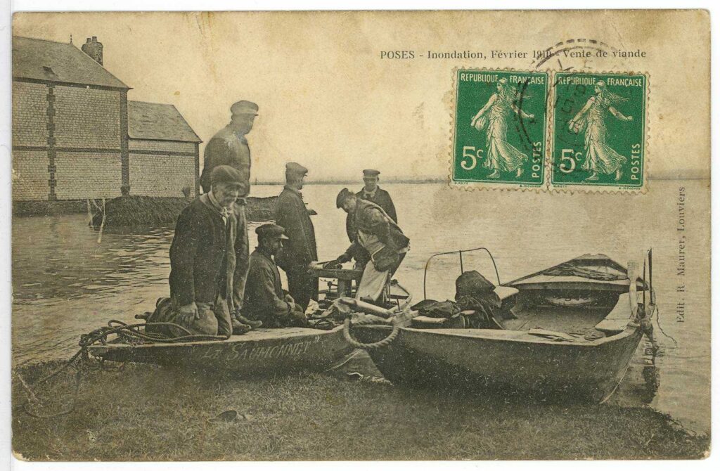 Carte postale 1910. Poses, inondation, février 1910, vente de viande. Embarcation dans la rue et personnage dessus (source : Archives Départementales 27)