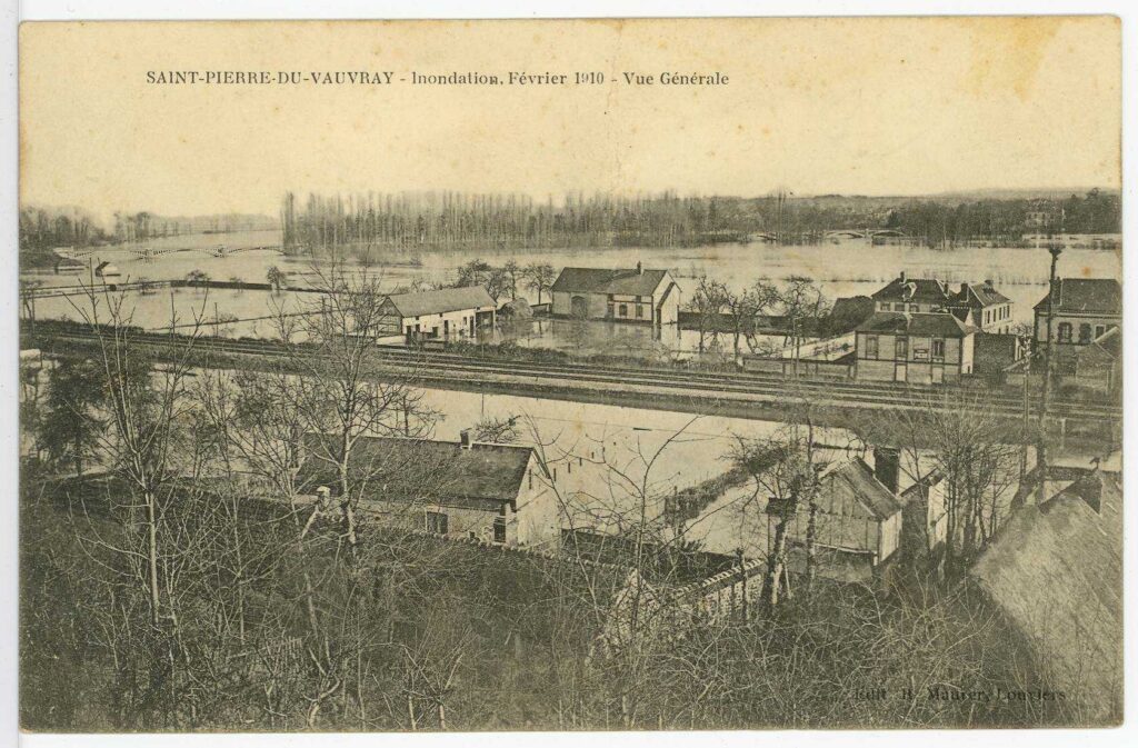 Carte postale 1910. Saint Pierre du Vauvray, pendant les inondations février 1910. Vue générale depuis le coteau avec les voies de chemin de fer et les habitations inondées. En arrière plan, le pont entre Andé et St Pierre du Vauvray et l'ïle du Bac ennoyée (source : Archives Départementales 27)