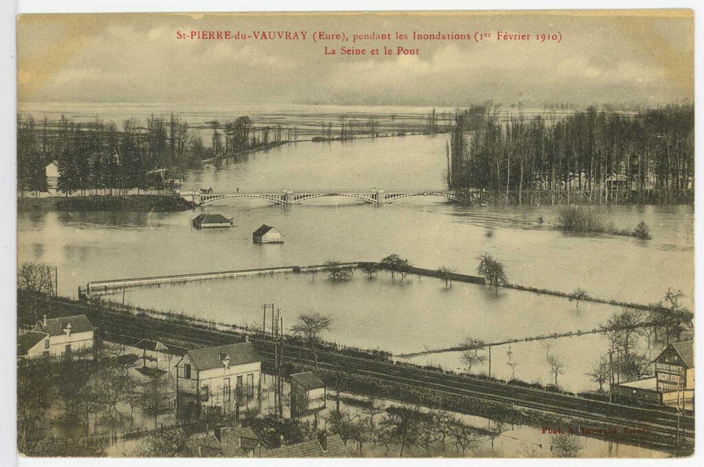 Carte postale 1910. Saint Pierre du Vauvray, pendant les inondations 1er février 1910. La Seine et le Pont. Vue de la rive gauche de la Seine inondée, avec des bâtiments entourés d'eau (source : Archives Départementales 27)
